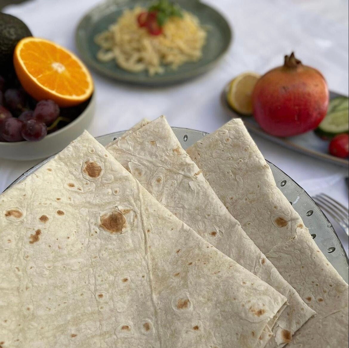 Folded köy yufkası on a glass dish, with orange, pomegranate and grapes behind it on a white tablecloth